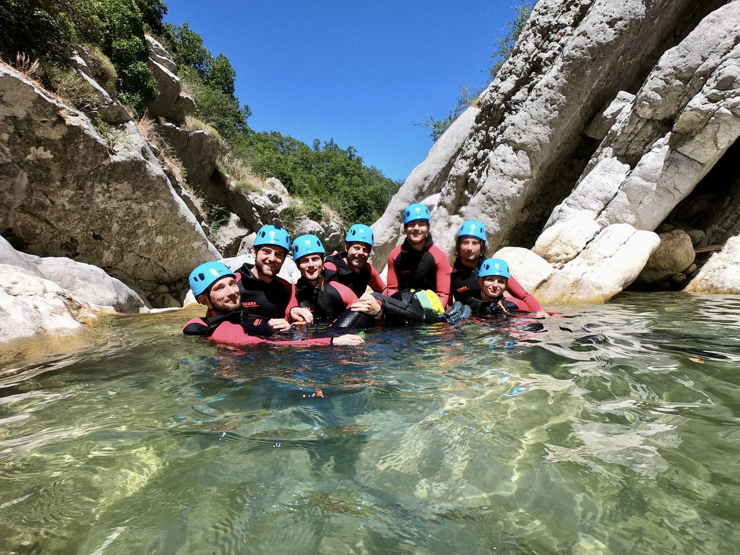 un groupe de participants à une sortie canyoning sourit dans une piscine naturelle d'eau translucide sous le ciel bleu de provence