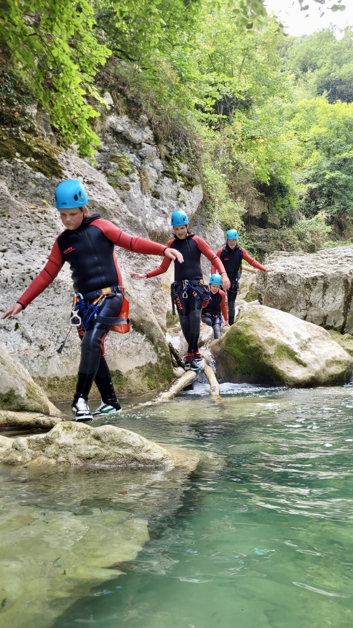 Un groupe d'enfants et d'adultes équipés de combinaisons et de casques bleus marchant en équilibre sur un tronc d'arbre au-dessus de l'eau.
