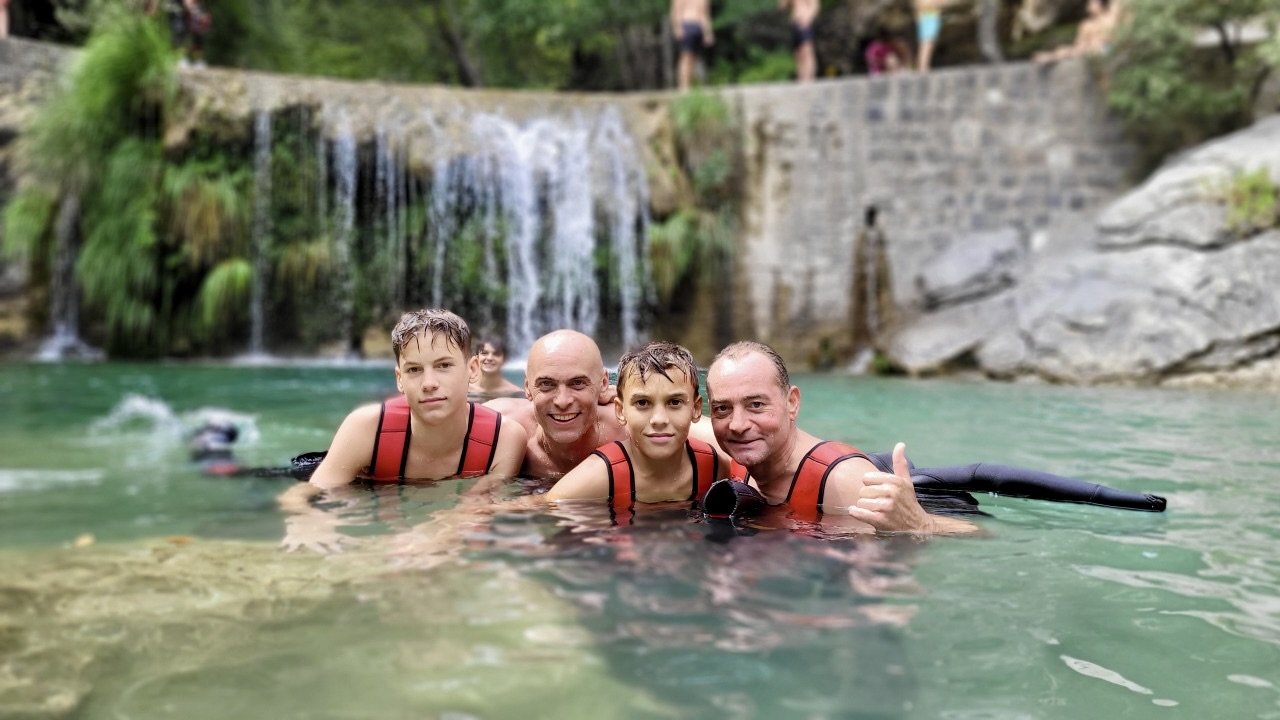 Portrait de quatre participants souriants dans l'eau translucide d'une vasque avec la cascade du canyon de la Barbaira en arrière-plan.